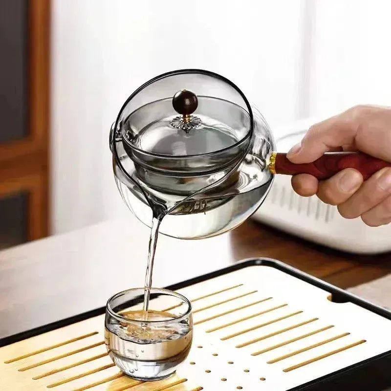 Transparent rotary glass teapot with wooden handle and built-in tea infuser on a table.