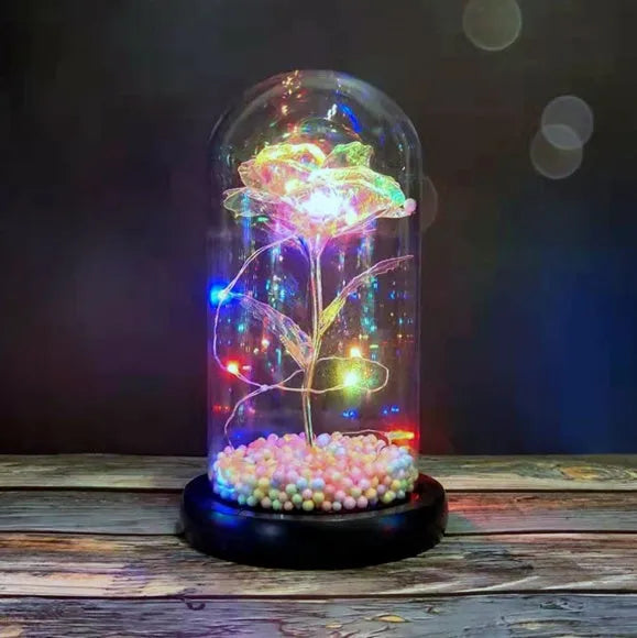 Artificial gold rose flower inside a glass lantern with LED lights and black base on a table.