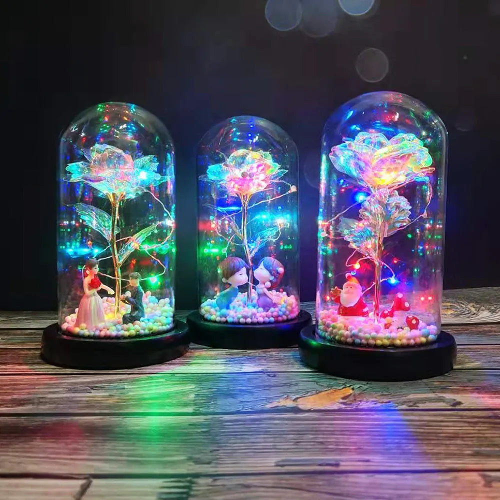 Artificial gold rose flower inside a glass lantern with LED lights and black base on a table.