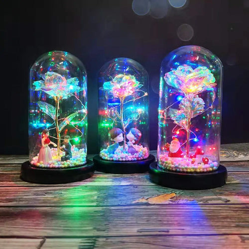 Artificial gold rose flower inside a glass lantern with LED lights and black base on a table.