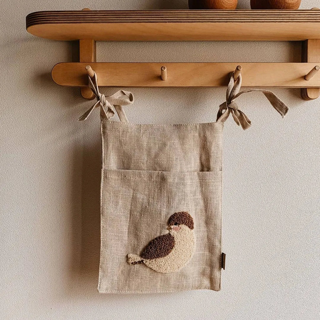 Khaki cotton linen hanging storage bag with multiple pockets mounted beside a bed.