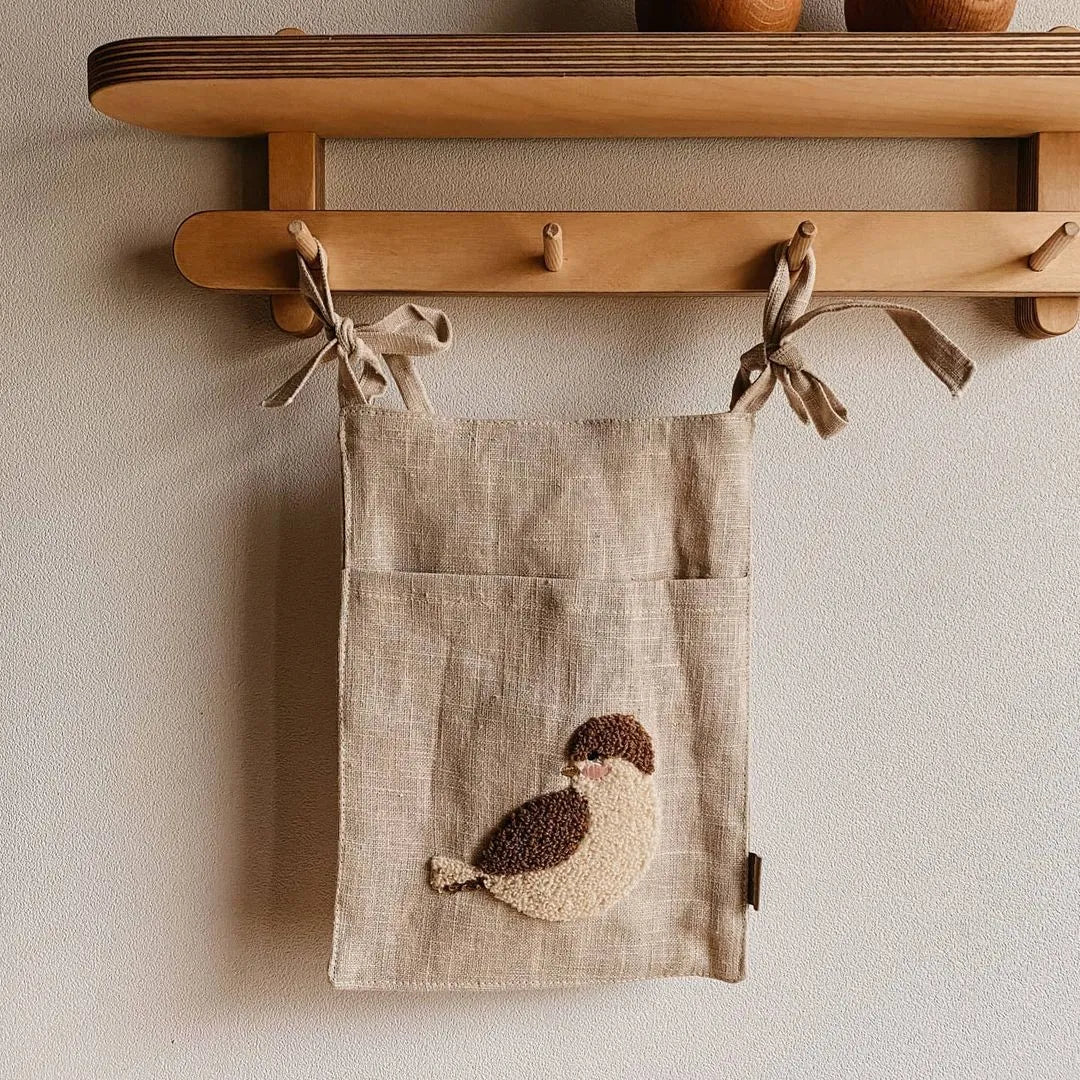 Khaki cotton linen hanging storage bag with multiple pockets mounted beside a bed.