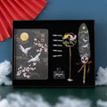 Chinese style metal feather pen with sheath and coloured feather collections next to a notebook.