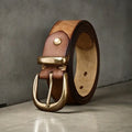 Men's black cattlehide leather jeans belt with a copper pin buckle on a wooden background.