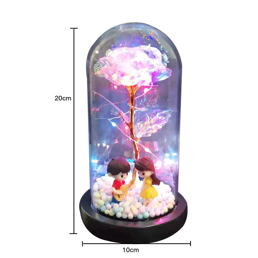 Artificial gold rose flower inside a glass lantern with LED lights and black base on a table.