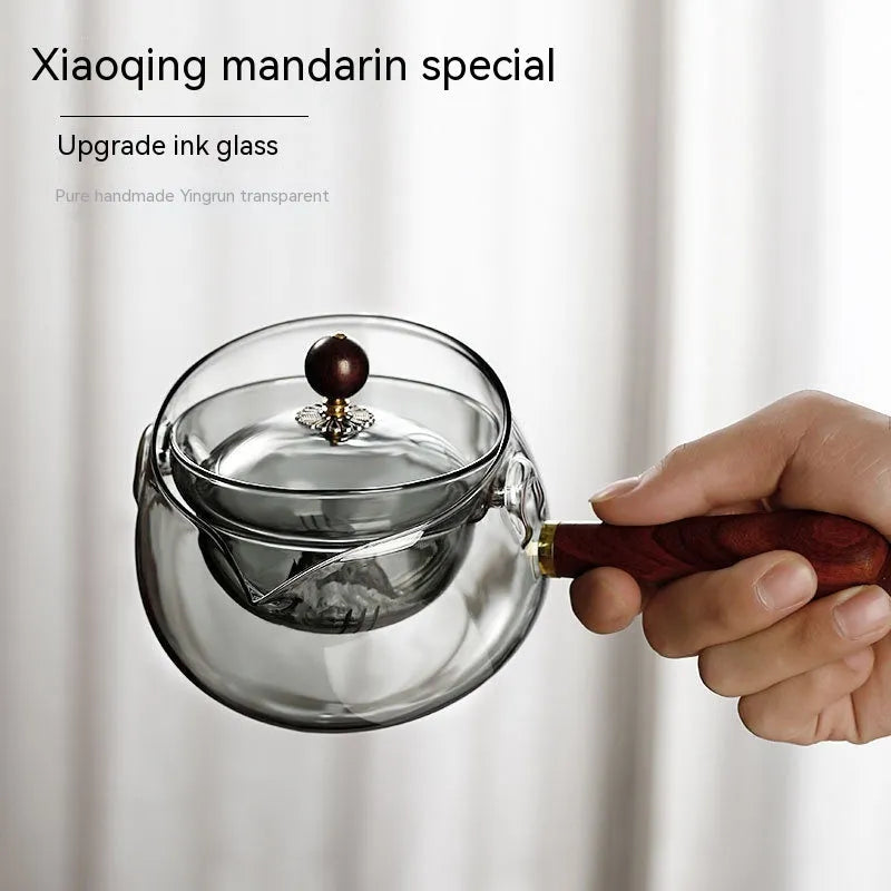 Transparent rotary glass teapot with wooden handle and built-in tea infuser on a table.