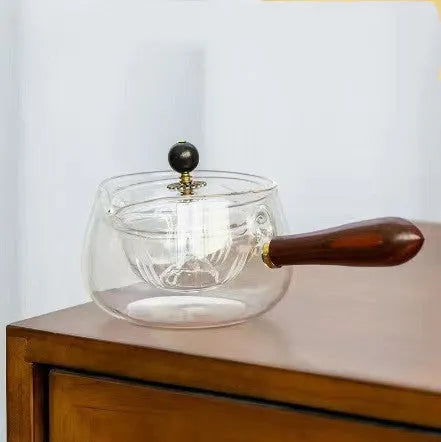 Transparent rotary glass teapot with wooden handle and built-in tea infuser on a table.