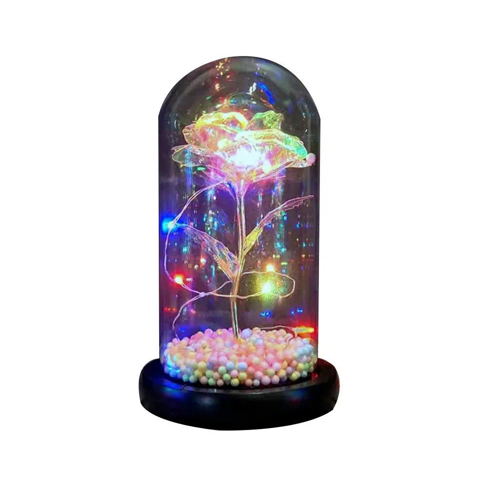 Artificial gold rose flower inside a glass lantern with LED lights and black base on a table.