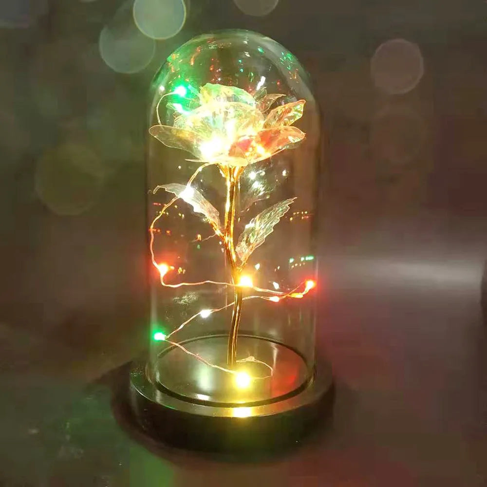 Artificial gold rose flower inside a glass lantern with LED lights and black base on a table.