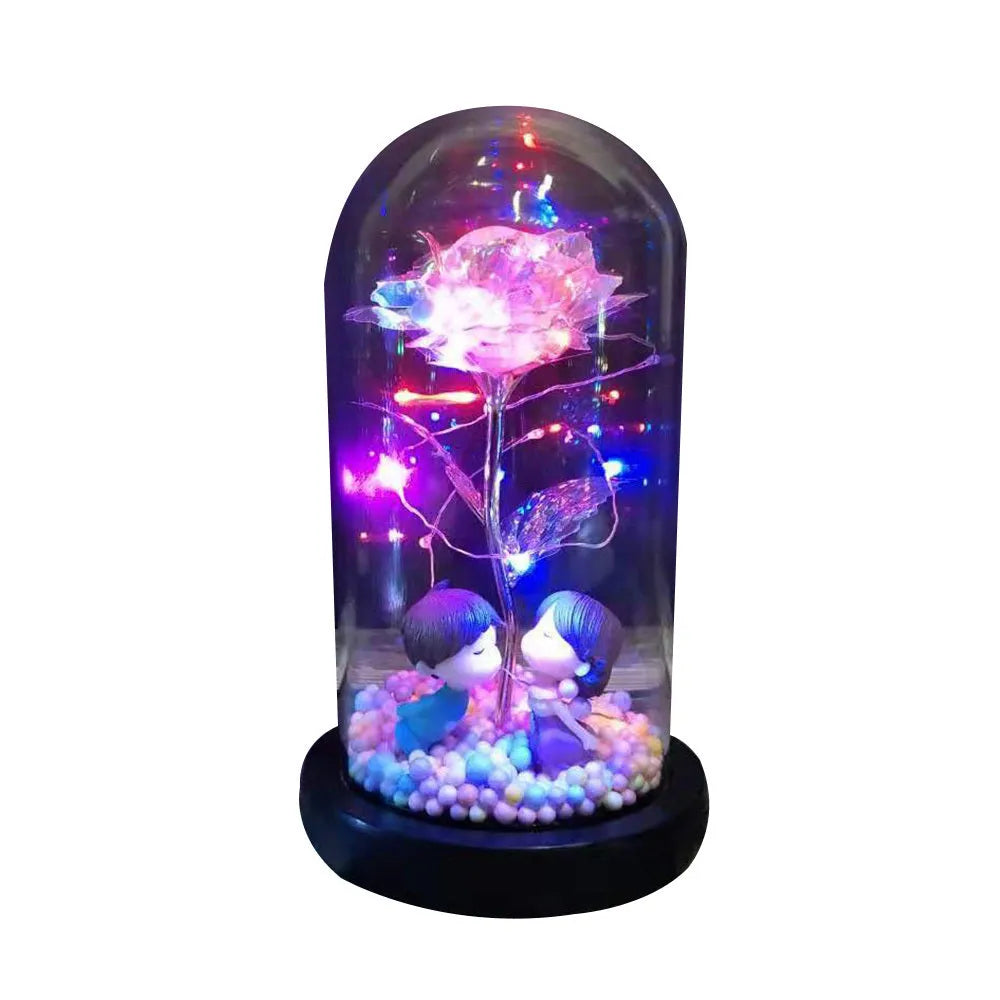 Artificial gold rose flower inside a glass lantern with LED lights and black base on a table.