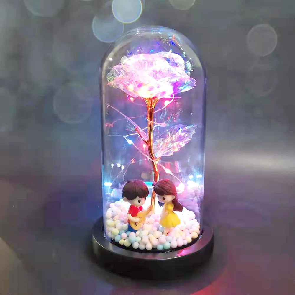 Artificial gold rose flower inside a glass lantern with LED lights and black base on a table.