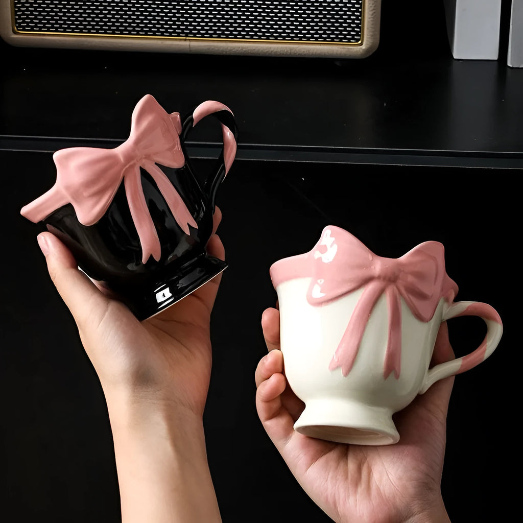 Cute white ceramic mug with a pink heart and black bow design, showing its handle.