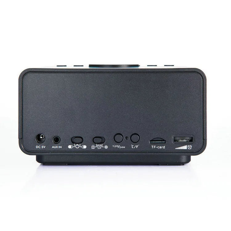 Black and white wireless alarm clock speaker with Bluetooth, radio, and USB playback functions on a bedside table.