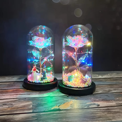 Artificial gold rose flower inside a glass lantern with LED lights and black base on a table.
