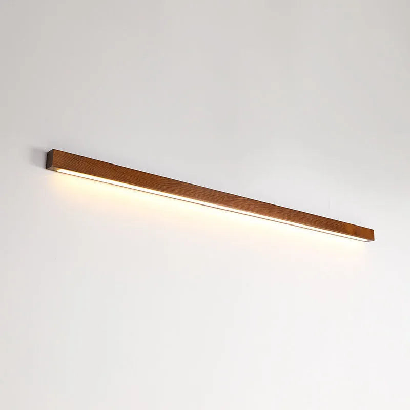Long, slim walnut-coloured wooden wall lamp with integrated LED light, shown in a minimalist room setting.