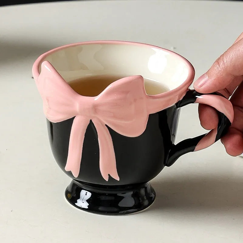 Cute white ceramic mug with a pink heart and black bow design, showing its handle.