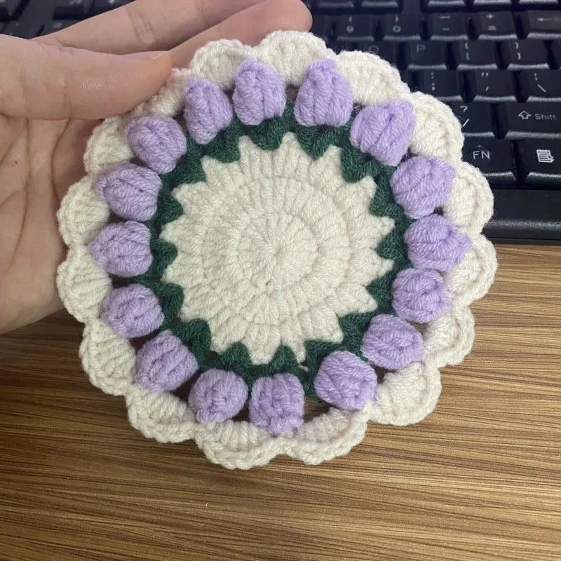 Set of four handmade woven floral coasters in pink, purple, red, and yellow on a wooden table.