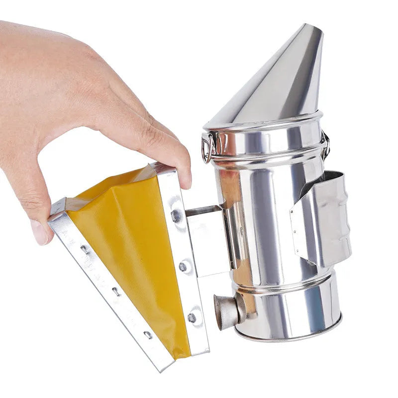 Stainless steel bee smoker with leather bellow on a wooden table against a blurred outdoor background