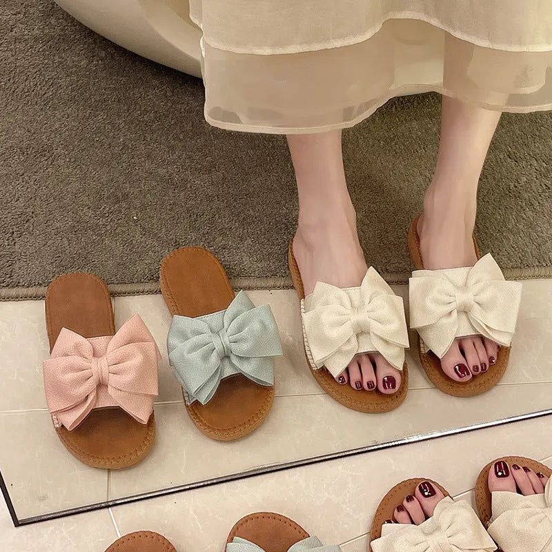 Women's beige slip-on suede sandals with a flat rubber sole and a small bow detail on the upper.