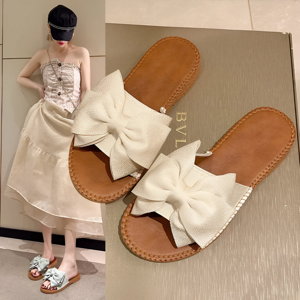 Women's beige slip-on suede sandals with a flat rubber sole and a small bow detail on the upper.