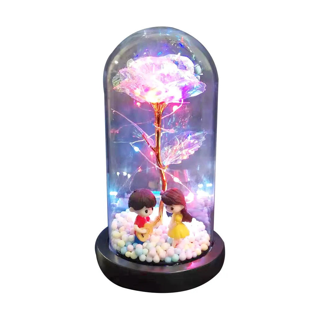 Artificial gold rose flower inside a glass lantern with LED lights and black base on a table.
