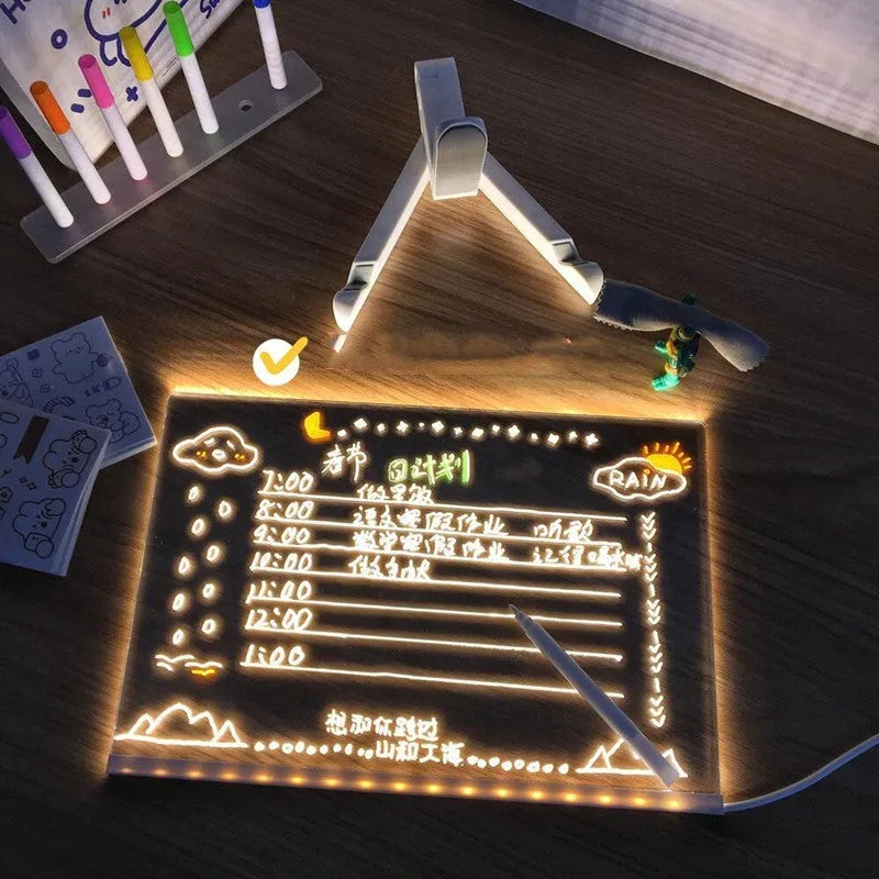 DIY acrylic LED message board lamp on a desk with colourful writing, showing USB cable and pens.