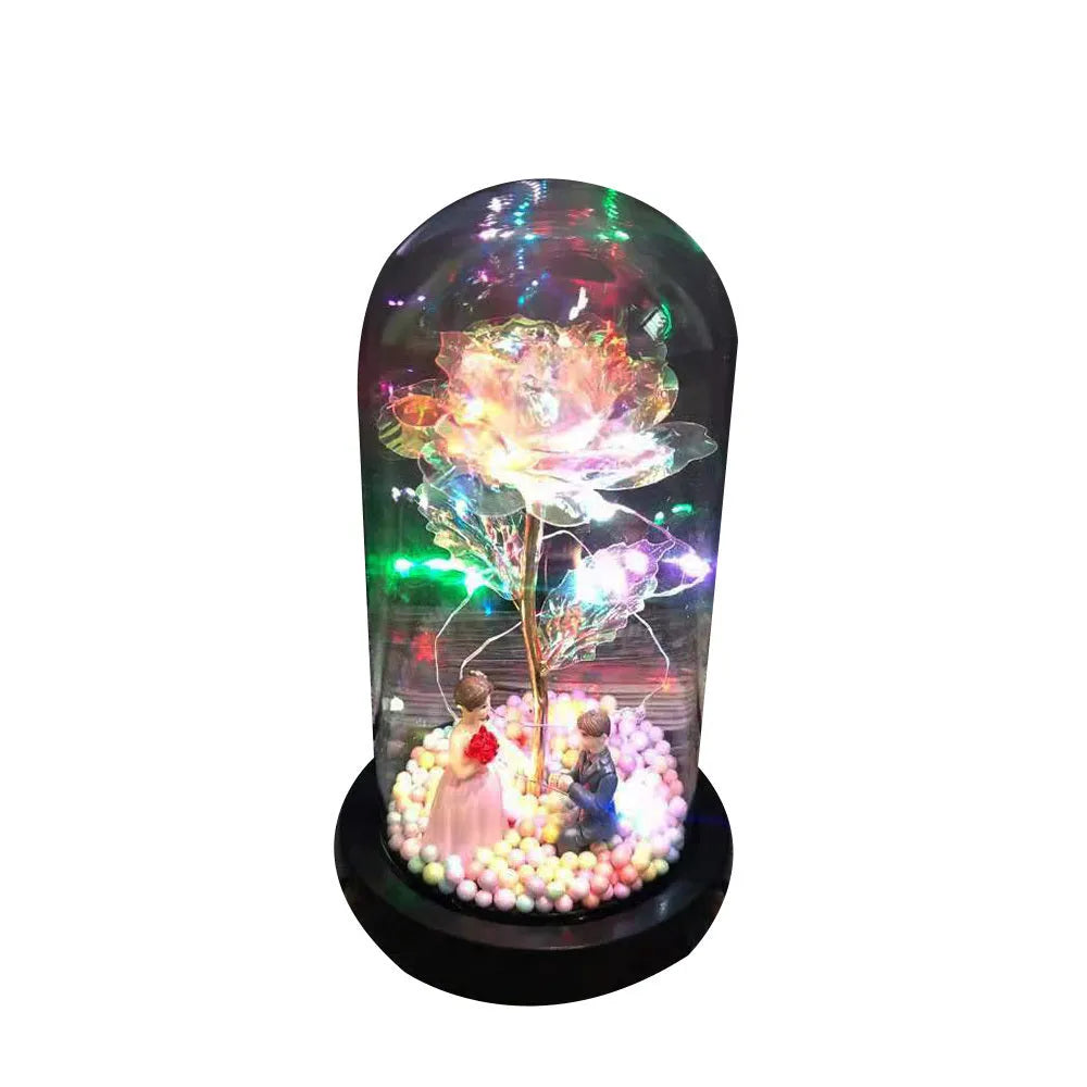 Artificial gold rose flower inside a glass lantern with LED lights and black base on a table.