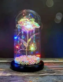 Artificial gold rose flower inside a glass lantern with LED lights and black base on a table.