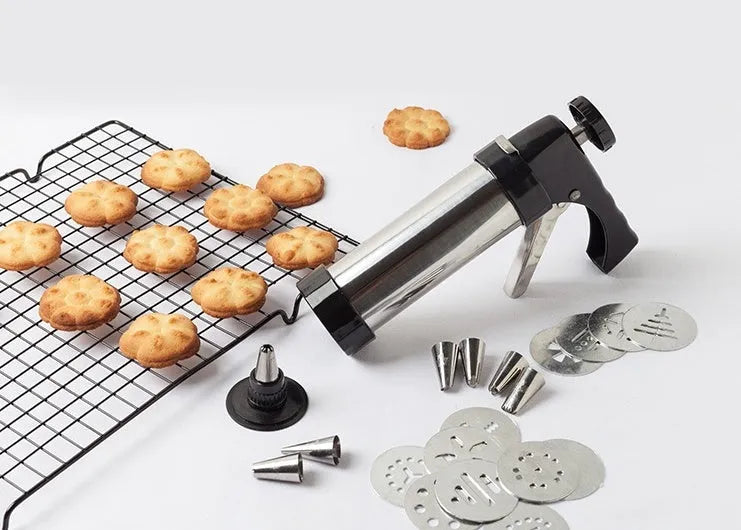 Stainless steel biscuit press gun with assorted cookie mould discs and icing nozzles on a baking tray.