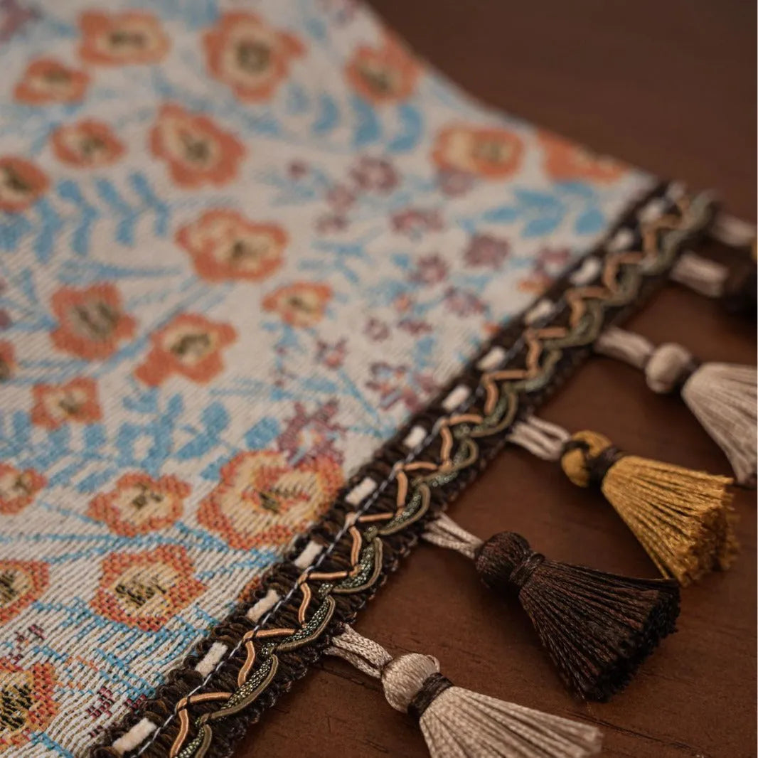 Beige jacquard fabric table runner with floral pattern and long tassels laid flat on a table.