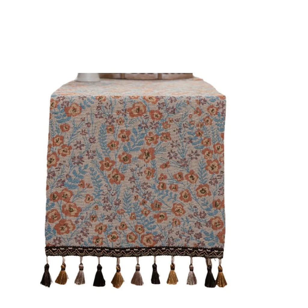Beige jacquard fabric table runner with floral pattern and long tassels laid flat on a table.