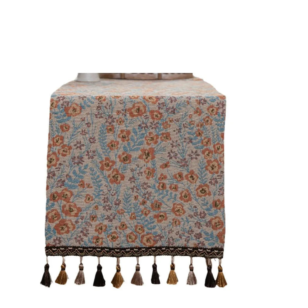 Beige jacquard fabric table runner with floral pattern and long tassels laid flat on a table.