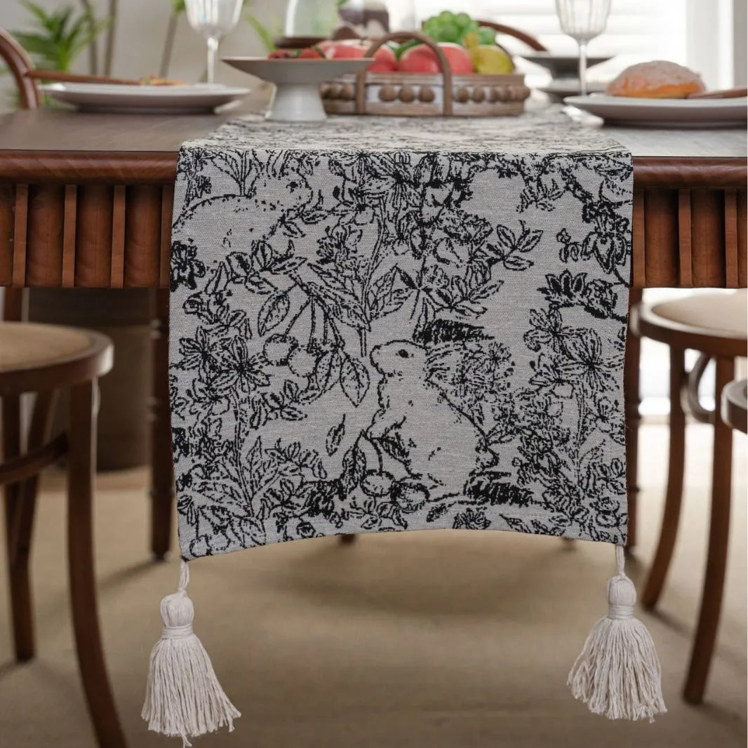 Beige jacquard fabric table runner with floral pattern and long tassels laid flat on a table.