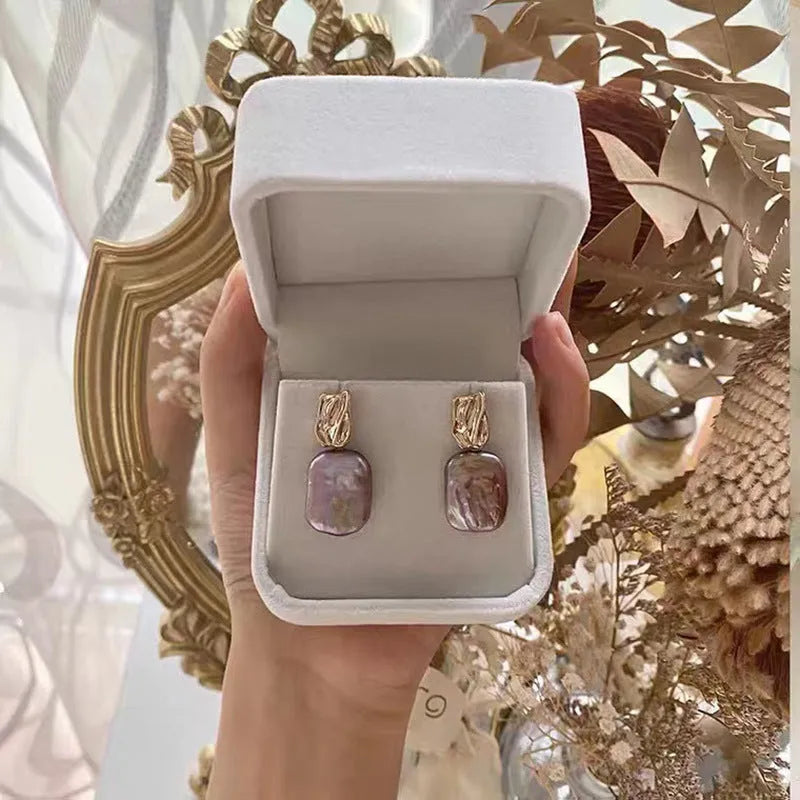 A pair of square stud earrings with a natural freshwater pearl set in a geometric alloy frame, shown in purple.