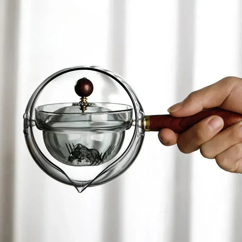 Transparent rotary glass teapot with wooden handle and built-in tea infuser on a table.