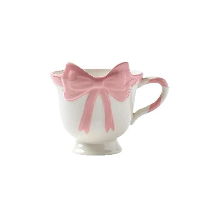 Cute white ceramic mug with a pink heart and black bow design, showing its handle.