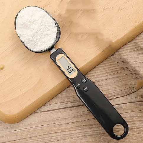 White digital kitchen scale with LCD screen displaying weight, shown next to a measuring spoon.