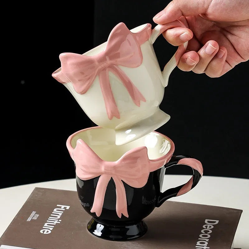 Cute white ceramic mug with a pink heart and black bow design, showing its handle.