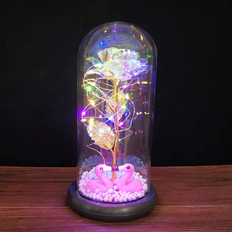 Artificial gold rose flower inside a glass lantern with LED lights and black base on a table.