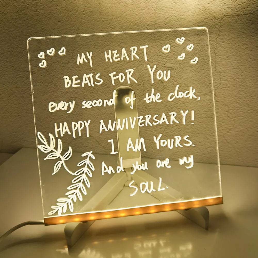 DIY acrylic LED message board lamp on a desk with colourful writing, showing USB cable and pens.