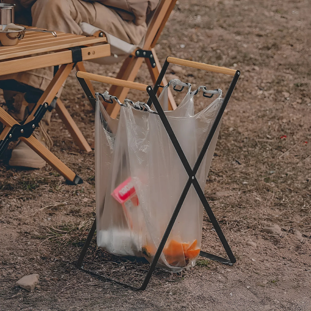 Black iron folding trash storage rack with a wooden handle for hanging bin bags.