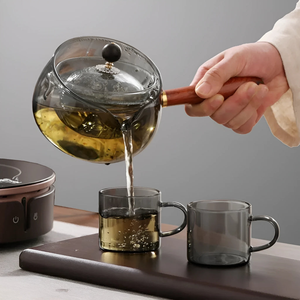 Transparent rotary glass teapot with wooden handle and built-in tea infuser on a table.