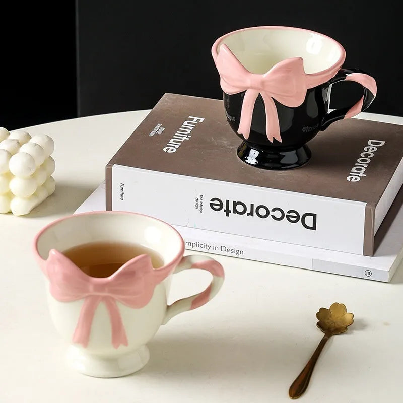 Cute white ceramic mug with a pink heart and black bow design, showing its handle.