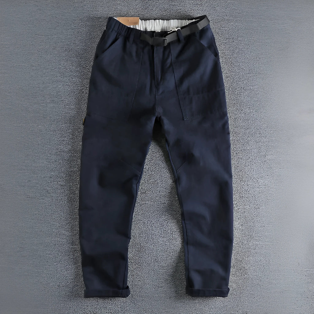 Man wearing khaki cotton work trousers with a buckle belt and structured three-dimensional cut.