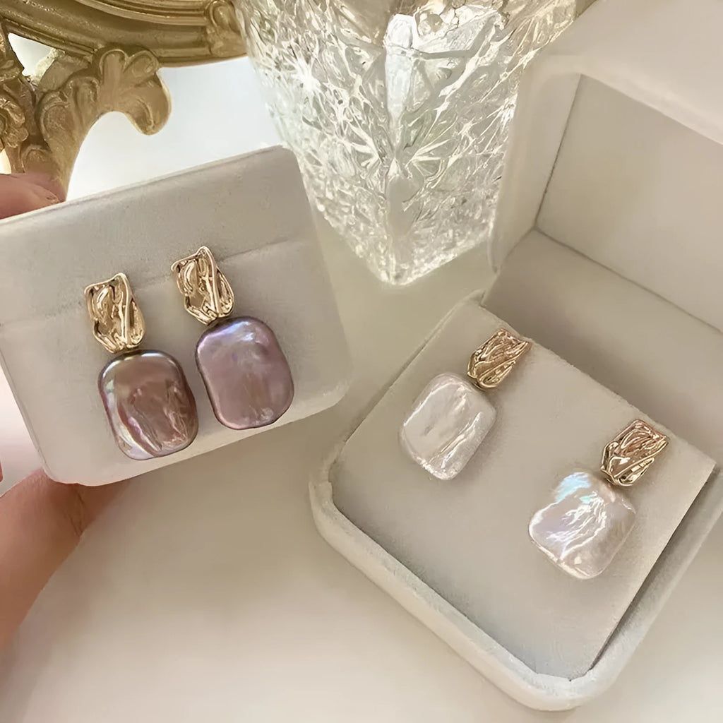 A pair of square stud earrings with a natural freshwater pearl set in a geometric alloy frame, shown in purple.