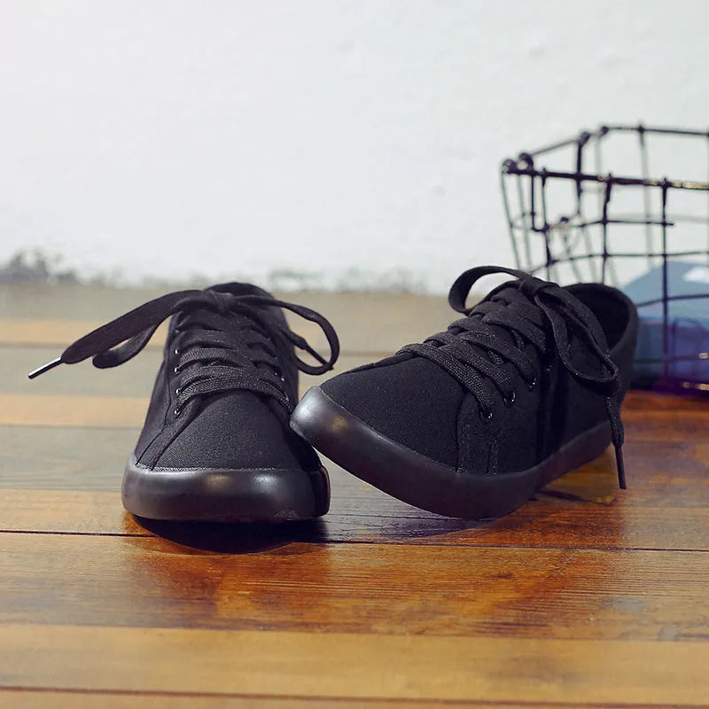 All black women's flat canvas shoes with white laces on a flat rubber sole.