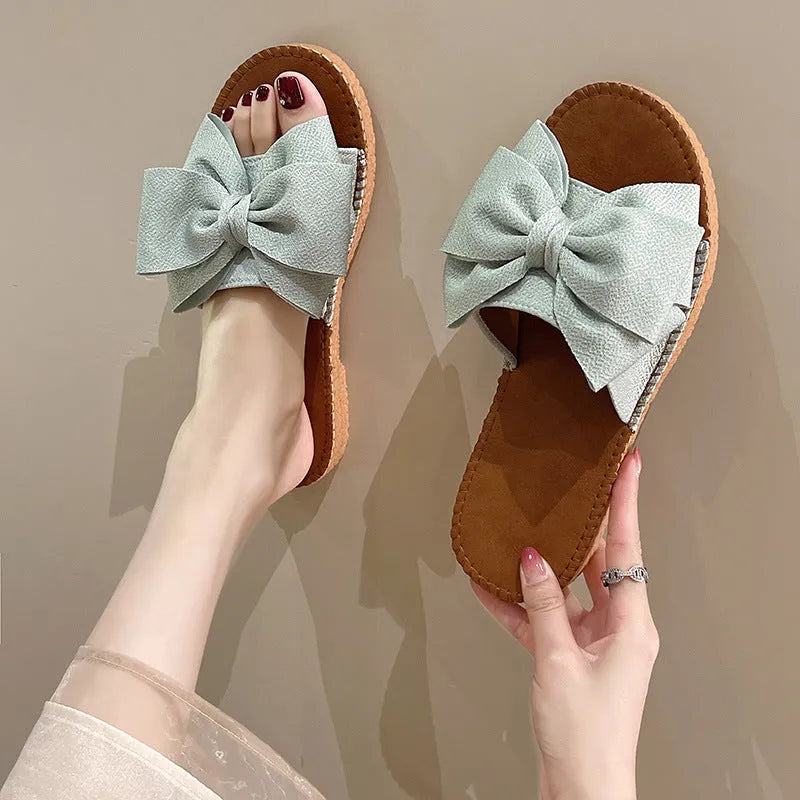 Women's beige slip-on suede sandals with a flat rubber sole and a small bow detail on the upper.