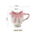 Cute white ceramic mug with a pink heart and black bow design, showing its handle.