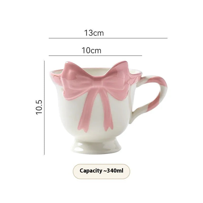 Cute white ceramic mug with a pink heart and black bow design, showing its handle.